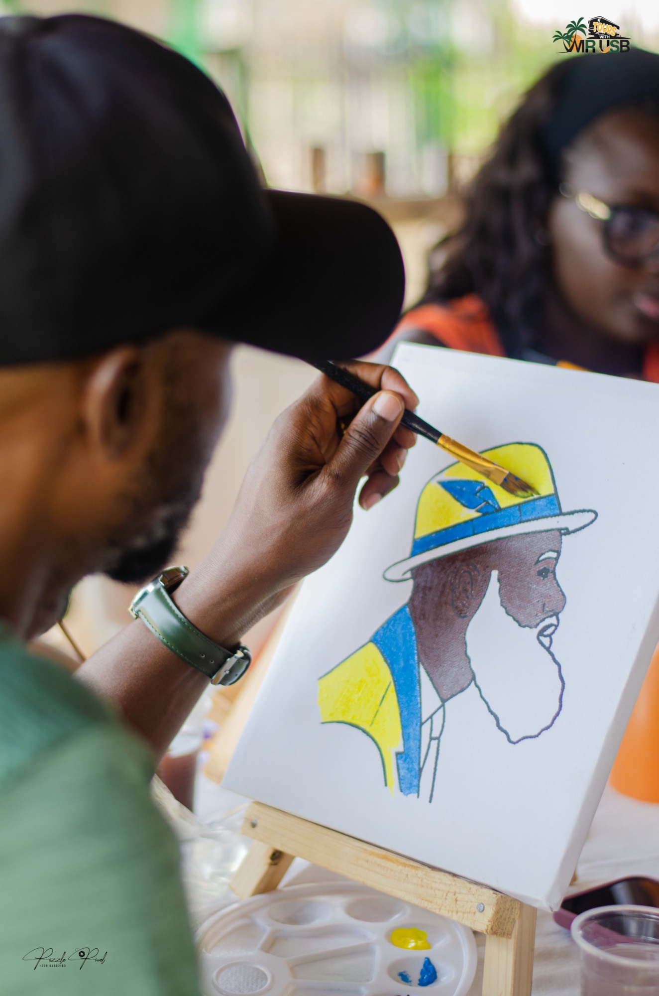A guest painting a portrait in blue and yellow, complete with a wide-brimmed hat